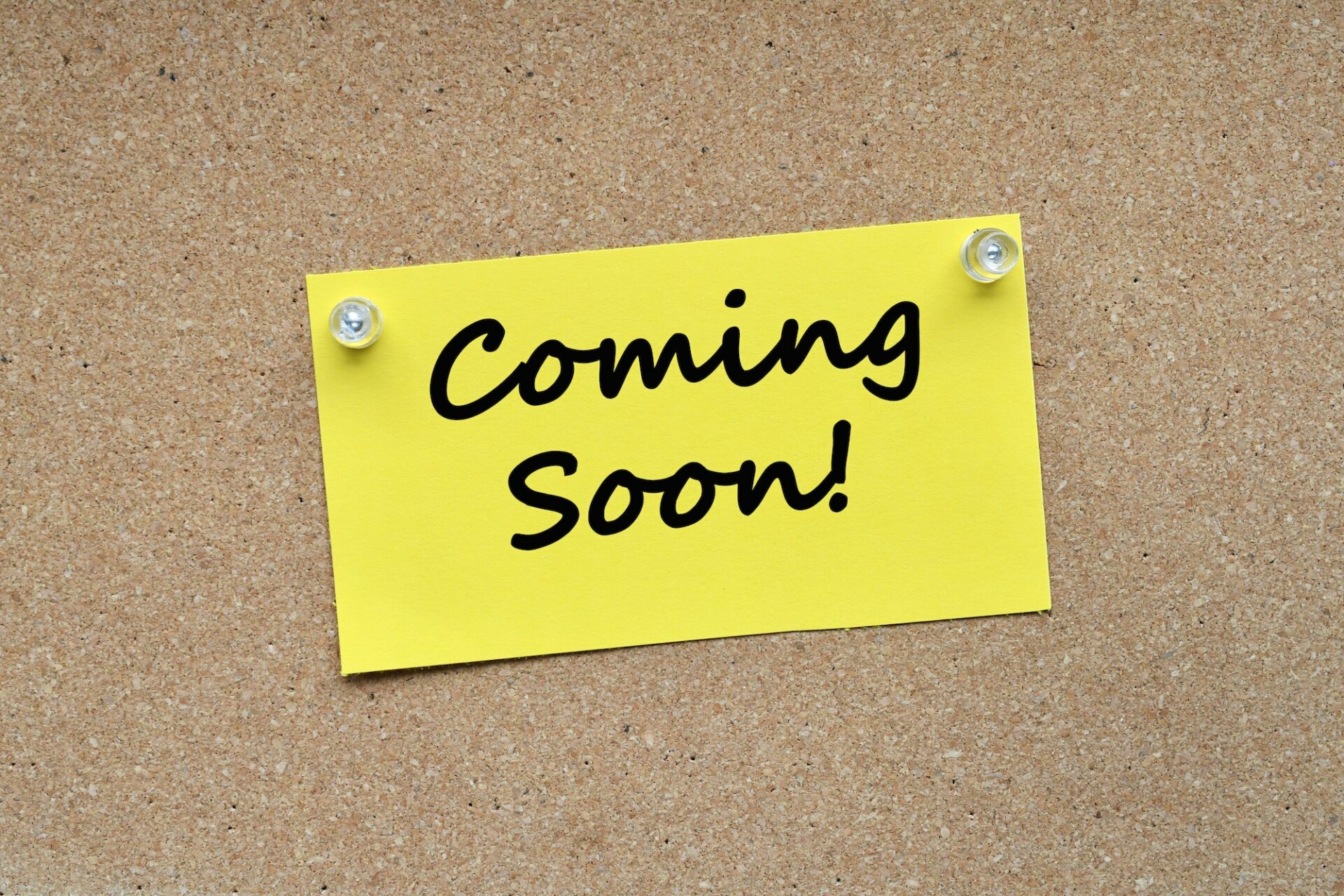 Coming Soon! on Yellow note card pinned with push pins on corkboard cork bulletin board.